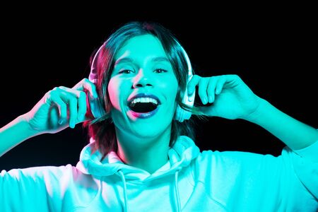 nightlife, technology and people concept - happy young woman in headphones wearing hoodie listening to music in neon lights over black backgroundの写真素材