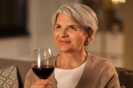 people, alcohol and drinks concept - happy senior woman drinking red wine from glass at home in eveningの写真素材