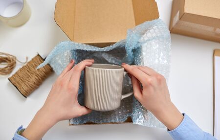 delivery, mail service, people and shipment concept - close up of female hands packing mug into...