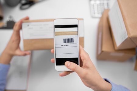 delivery, mail service, people and shipment concept - close up of woman's hands with smartphone scanning barcode on parcel box at post officeの写真素材
