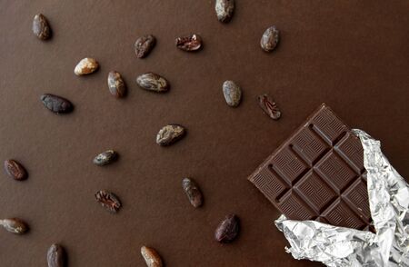 sweets, confectionery and food concept - dark chocolate bar in foil wrapper and cocoa beans on brown...