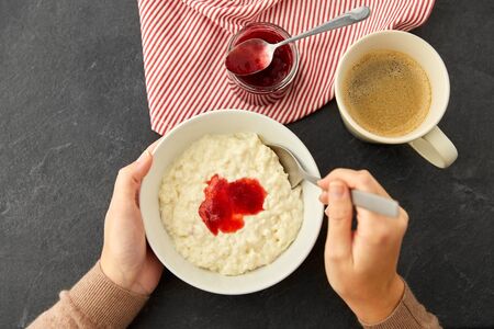 food and breakfast concept - hands of woman with spoon eating porridge in bowl with fruit jam and...