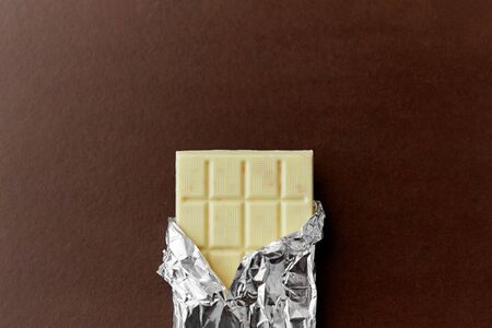 sweets, confectionery and food concept - white chocolate bar in foil wrapper on brown backgroundの写真素材