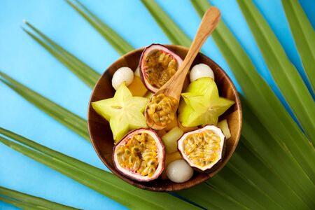 food, summer and healthy eating concept - mix of different exotic fruits in wooden plate with spoon and green fan palm leaf on blue backgroundの写真素材