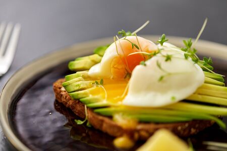 food, eating and breakfast concept - toast bread with sliced avocado, pouched egg and greens on...
