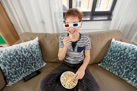 people and childhood concept - happy smiling boy in 3d movie glasses eating popcorn and watching tv at homeの写真素材