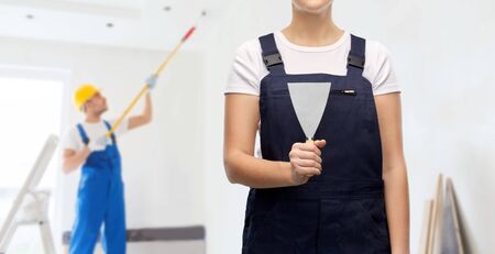repair, construction and building concept - close up of female plasterer or builder with putty knife over painter painting ceiling backgroundの写真素材