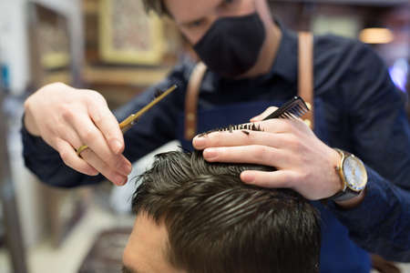 grooming, hairdressing and health safety concept - close up of male client and hairdresser wearing face medical mask for protection from virus disease with comb and scissors cutting hair at barbershopの写真素材
