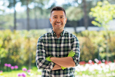 gardening and people concept - portrait of happy smiling middle-aged man with crossed arms in gloves at summer gardenの写真素材