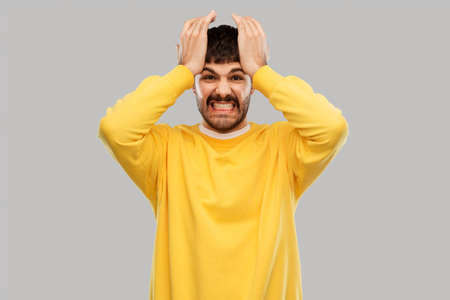 stress, emotions and people concept - angry young man in yellow sweatshirt holding to head over grey...