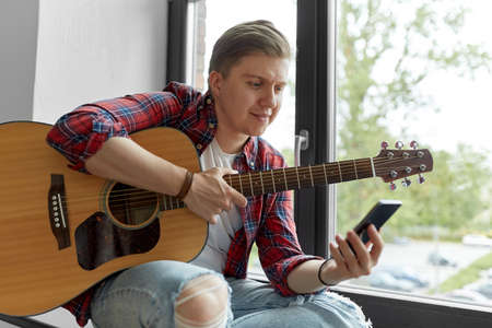 young man playing guitar sitting on windowsill