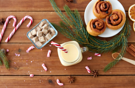 christmas and seasonal drinks concept - eggnog in glass mug with candy cane decoration, cinnamon buns, fir tree brunch and aromatic spices on wooden backgroundの写真素材