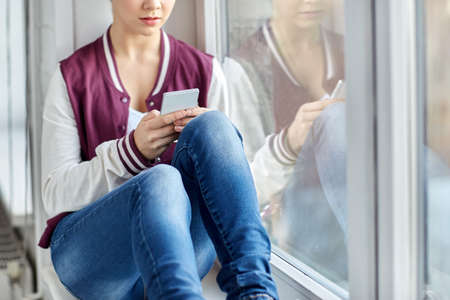 teenage girl with smartphone sitting on windowsill