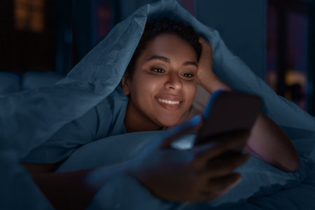 woman with smartphone under duvet in bed at nightの写真素材