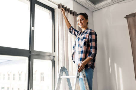 woman on ladder hanging curtains at home