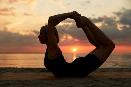 woman doing yoga bow pose on beach over sunset