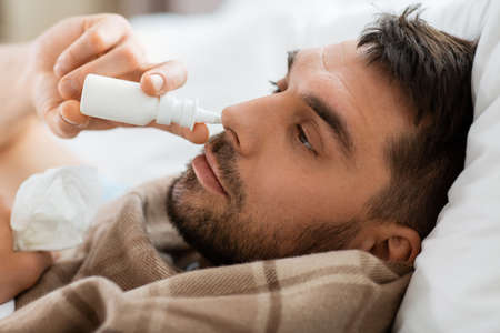 sick man with nasal spray lying in bed at homeの写真素材