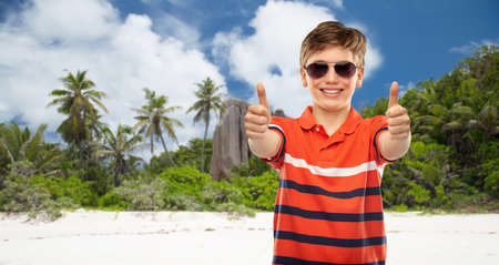 happy boy in sunglasses showing thumbs up on beachの写真素材