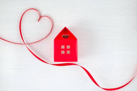 Red ribbon heart on a white wooden background, red house with a heart shape window in a romantic, valentine, family or wedding concept. Empty space for your text.の写真素材