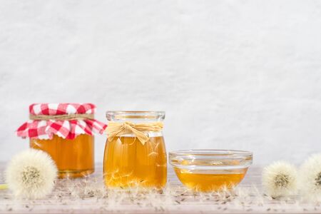 Dandelion jam, honey, jelly in a glass jar on a wooden table, white background with fresh flowers, dandelion airy seed heads, seeds, blow balls. Medicine, healthy food, health benefits from nature.の写真素材