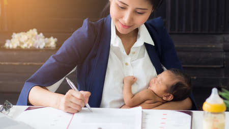 Business woman holding baby in her arms while working, signing contract. Single mum taking care of newborn in the office. Can use for varieties of concepts.の写真素材