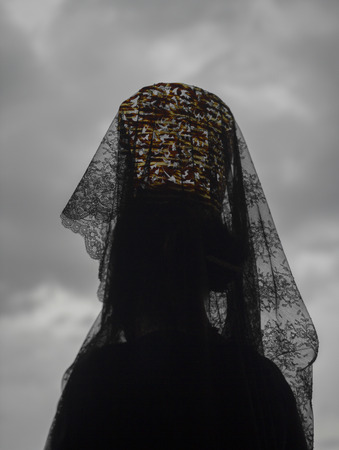 Woman wearing a headdress of black Spanish lace lifted high on a traditional comb standing with her back to the camera agaisnt a cloudy grey sky during a Holy Week in Andalucia, Spainの写真素材
