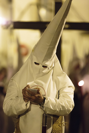Religious Catholic penitent dressed in a conical hat and robes standing writing notes during the Holy Week procession in Seville, Spainのeditorial素材