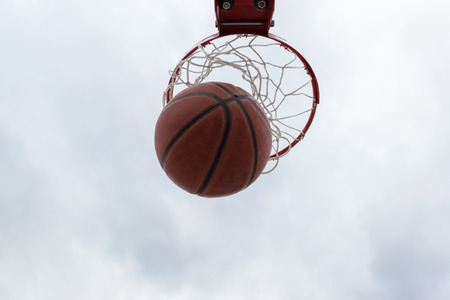 Ball inside red basketball hoop, basket against cloudy sky. Outdoor basketball courtの写真素材