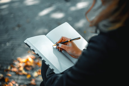 Young woman writing in a diary in an autumn park. Closeup image of a woman writing on a blank notebook. Woman working outdoors.の写真素材