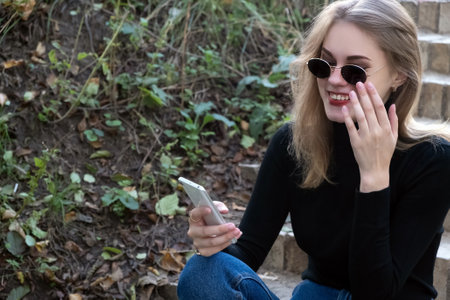 Young happy woman in black turtleneck looks into mobile phone touching her sunglasses with her hand...