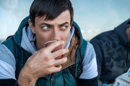 Serious brunette man drinking coffee or tea from disposable paper cup outdoors