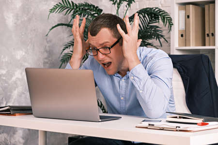 Portrait of an indignant man in a shirt and glasses, looking at the laptop screen with his hands up, shouting at the monitor, sitting in the office against the backdrop of a rack with folders.の写真素材