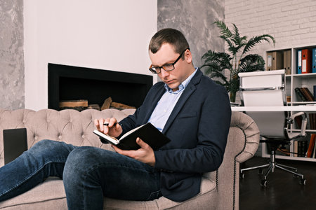 Young successful man in suit and glasses works with notebook or planner while sitting in spacious office on couch against background of fireplace. Concept of success, business and time managementの写真素材