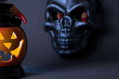Halloween background. A close-up of glowing pumpkin with a carved face, with human scary fingers on top, with a black mask skull in the background, with fingers sticking out of the eye socketsの写真素材