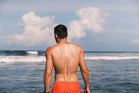 Rear view of a sporty man in shorts standing in the ocean against the backdrop of waves on a sunny...