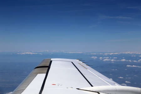 Wing of the plane on blue sky background and snowy mountains below, view from window of a jet plane wing with beautiful weatherの写真素材