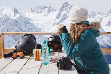 A girl with pleasure drinks tea in a village cafe in the background of snow mountains. A woman snowboarder relaxes outdoors. Winter peaks in clear weather. Calm Alpine look, lifestyle sport, travel.の写真素材