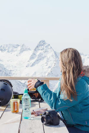 A girl with pleasure drinks tea in a village cafe in the background of snow mountains. A woman snowboarder relaxes outdoors. Winter peaks in clear weather. Calm Alpine look, lifestyle sport, travel.の写真素材