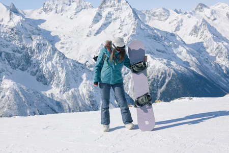 A snowboarder girl in the background of the mountains in the winter season enjoys the scenery. Vacation in the Alpine ski resort. The concept of snowboarding, success, prosperity, lifestyle, sport, recreation, adventure, travel.の写真素材