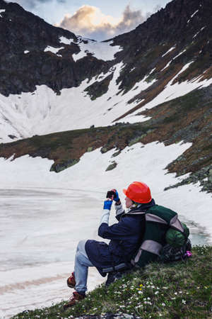A girl with a backpack in the mountains on the shore of the snowy lake and takes pictures of the peaks with her smartphone. Concept of a lifestyle hipster traveler, adventure, free tourism.の写真素材