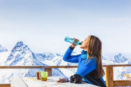 Woman traveler drinks water from a bottle in a cafe in a ski resort sunbathing in the snow at the top of the mountain. Travel and Adventure lifestyle concept. Stunning winter alpine landscape.の写真素材