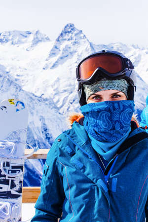 Girl with ski mask and snowboard on the background of snow-capped mountains enjoying a sunny winter...