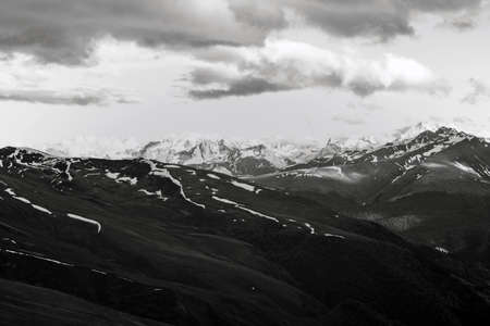 Monochrome black and white view of high slopes of hills and mountains covered with snow overcast on a spring day with sailing past the clouds. Concept of weather phenomenon and anticycloneの写真素材