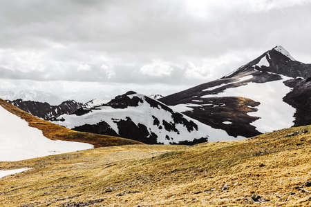 Beautiful view of a green hill from high snow-capped mountains against a cloudy sky and gray clouds breaking through them with sunlightの写真素材