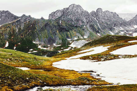Stunning picturesque view of the green-yellow hills covered with snow against the background of high gray rocks and overcast sky. Pristine nature conceptの写真素材