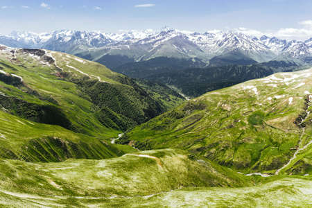 Stunning beautiful fabulous view of green hills and grasses against the high snow-covered rocks of the blue sky and clouds. Ð¡oncept of dventure and hiking in inaccessible clean places.の写真素材