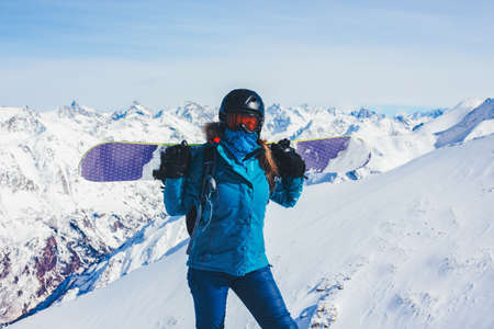 Sports background snowboarder girl in helmet and mask to high mountains, holds a snowboard on his shoulders. Lifestyle winter recreation and extreme adventure concept, activities vacations outdoor.の写真素材