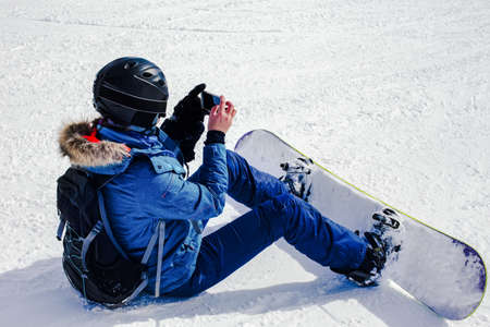 Shot female snowboarder in helmet and mask using smartphone, while riding snowboarding taking photos of winter nature in the mountains. Connectivity mobility lifestyle technology. Sports and adventureの写真素材