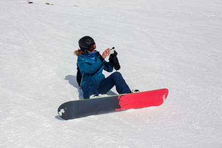 Girl snowboarder in helmet and mask takes pictures using smartphone, while riding snowboarding. Connectivity mobility lifestyle technology. Travel Lifestyle sport and extreme, adventure activities.の写真素材
