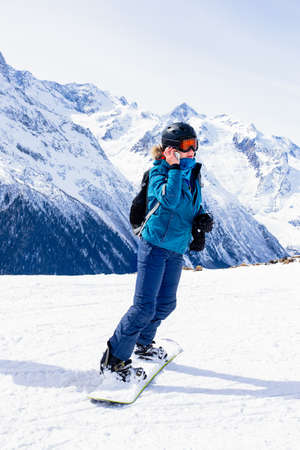 Girl snowboarder in helmet and mask talking on smartphone. Connectivity mobility technology. Travel Lifestyle sport and extreme, adventure activities. Ski resort in the mountains vacations.の写真素材